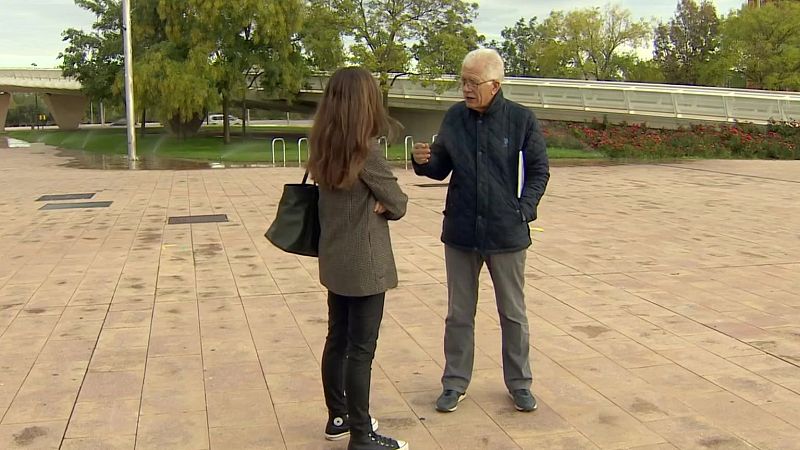 Dos personas conversan en una plaza pavimentada, con una zona ajardinada al fondo bajo un cielo cubierto.