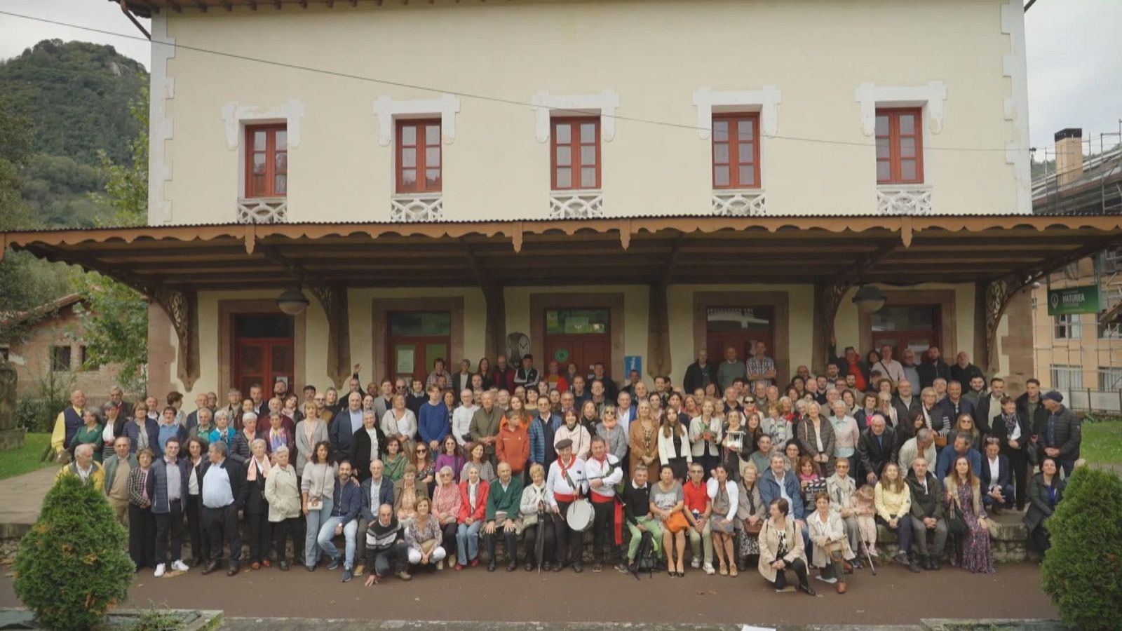 Numeroso grupo de personas posa frente a un edificio histórico de dos plantas con marquesina de madera, destacando dos hombres con traje tradicional y tambor.