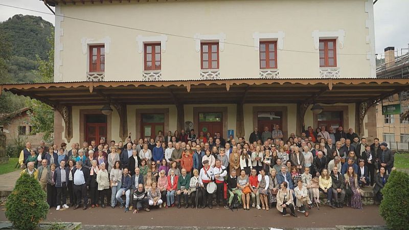 Numeroso grupo de personas posa frente a un edificio histórico de dos plantas con marquesina de madera, destacando dos hombres con traje tradicional y tambor.