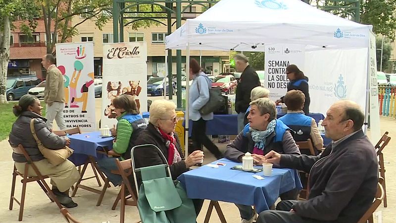 Personas mayores conversan sentadas en mesas con manteles azules bajo una carpa blanca durante una actividad comunitaria al aire libre.
