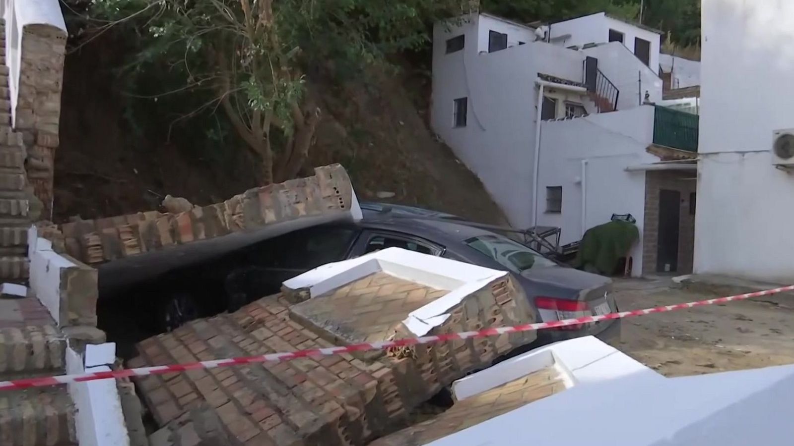 Un muro de ladrillo colapsa sobre un turismo gris en una zona residencial con viviendas escalonadas, delimitado por cinta policial.