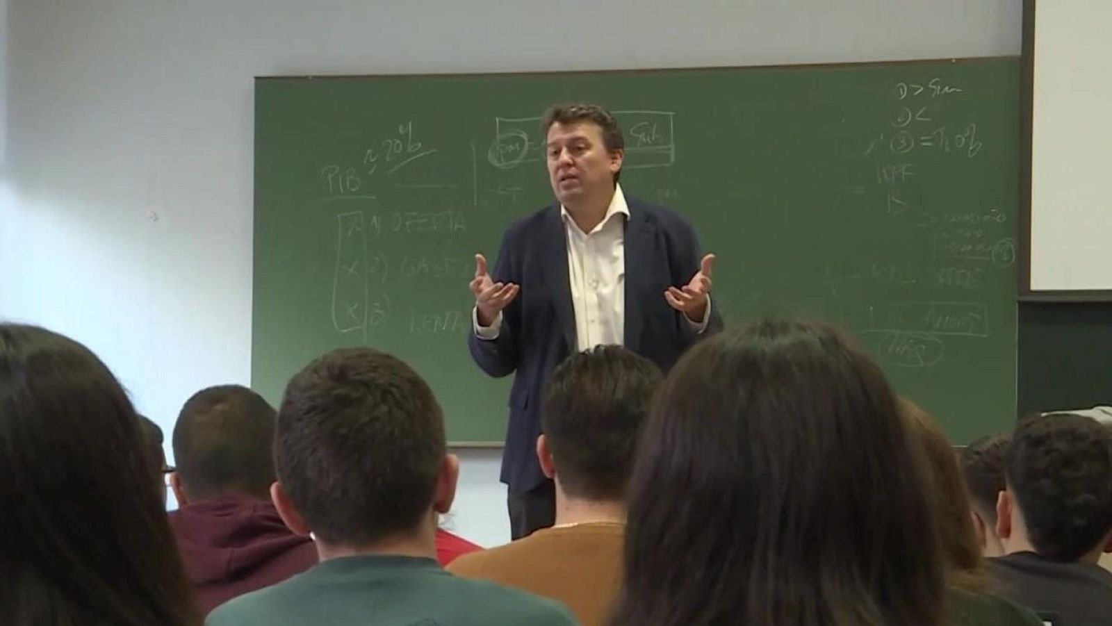 Juan Bravo imparte una clase ante estudiantes frente a una pizarra con fórmulas y esquemas económicos escritos con tiza blanca.