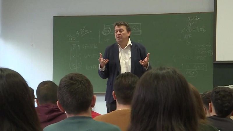 Juan Bravo imparte una clase ante estudiantes frente a una pizarra con fórmulas y esquemas económicos escritos con tiza blanca.