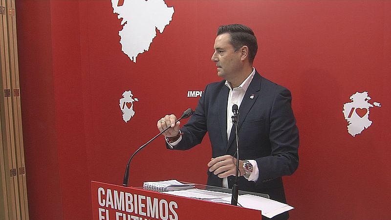 Hombre de mediana edad con traje azul y camisa blanca comparece ante un atril rojo con un lema escrito, ajustando un micrófono en una sala con fondo corporativo rojo.