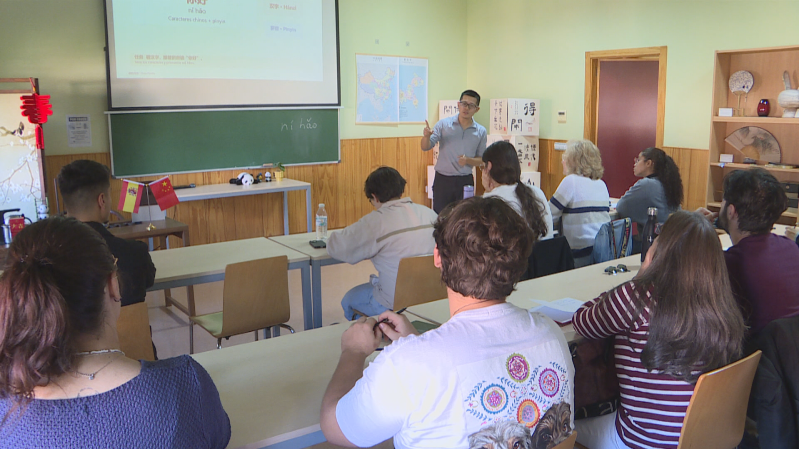Profesor impartiendo clase de cultura china frente a una pizarra con texto en pinyin, banderas de España y China sobre la mesa, y estudiantes prestando atención.
