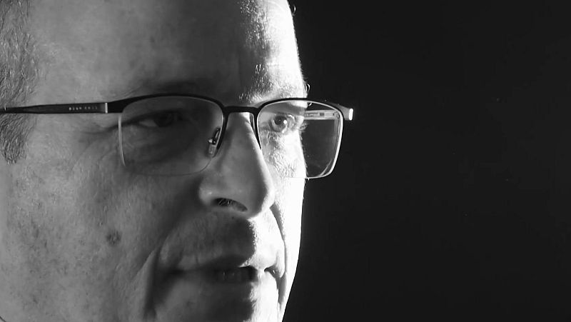 Primer plano en blanco y negro de un hombre serio con gafas y cabello entrecano, iluminado con una dramática luz lateral sobre fondo oscuro.