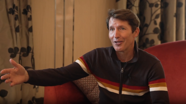 Plano medio de James Blunt sentado en una butaca roja, vistiendo chaqueta marrón y hablando de forma animada.