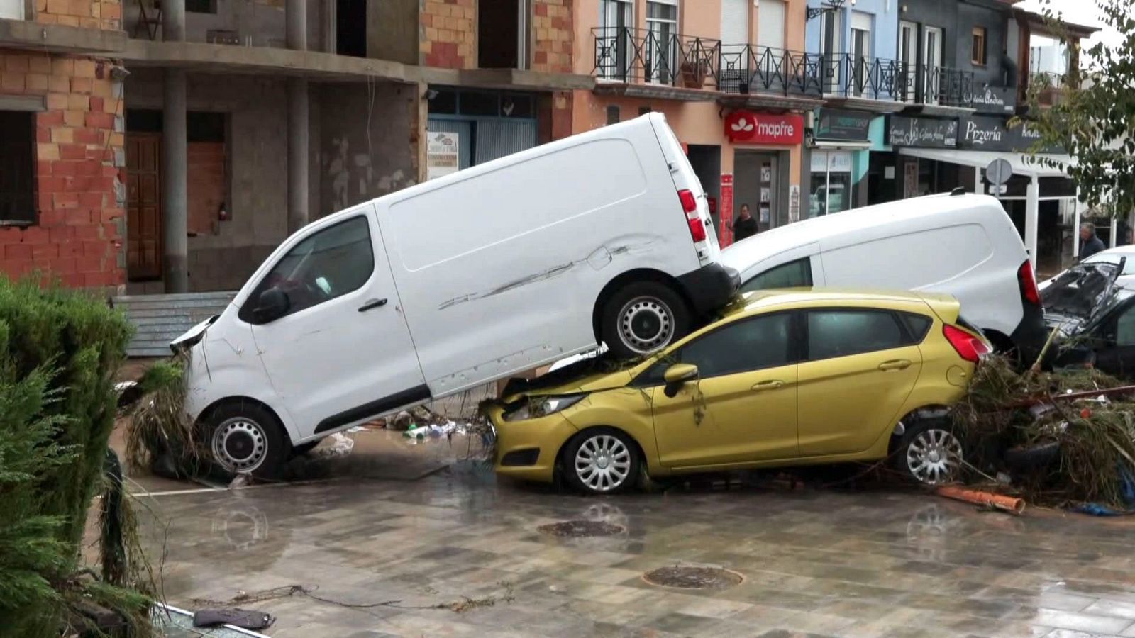Furgoneta blanca volcada sobre un coche amarillo y rodeada de barro y ramas en una calle tras una inundación.