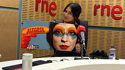 La cantante Anni B Sweet sostiene la portada de su disco 'Alegr�a' en un estudio de radio, sentada ante una mesa con un micr�fono de RNE y paneles de madera al fondo.