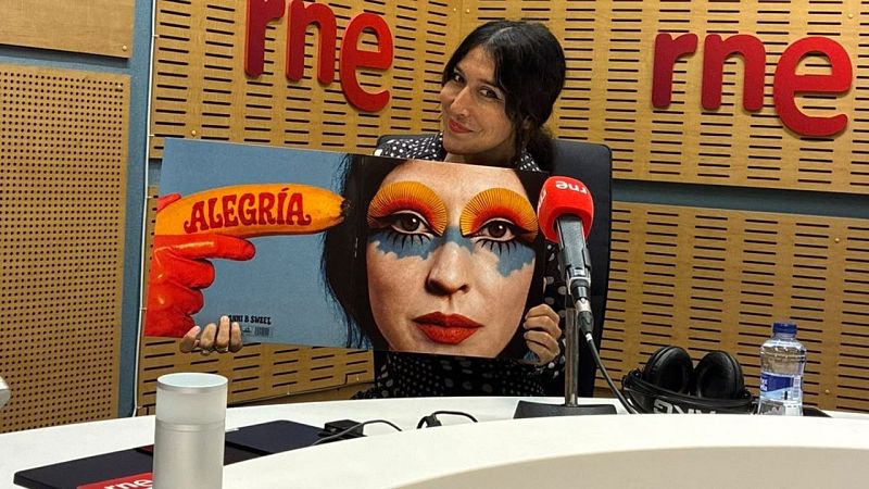 La cantante Anni B Sweet sostiene la portada de su disco 'Alegría' en un estudio de radio, sentada ante una mesa con un micrófono de RNE y paneles de madera al fondo.