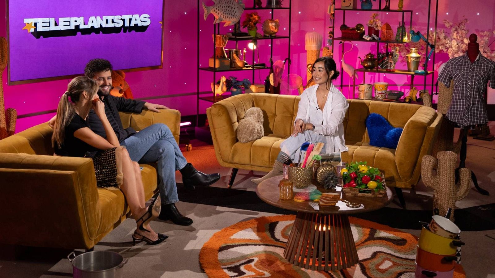 Plató de televisión iluminado en tonos fucsia con dos presentadores y la invitada Marina Rivers sentados en sofás de terciopelo mostaza.