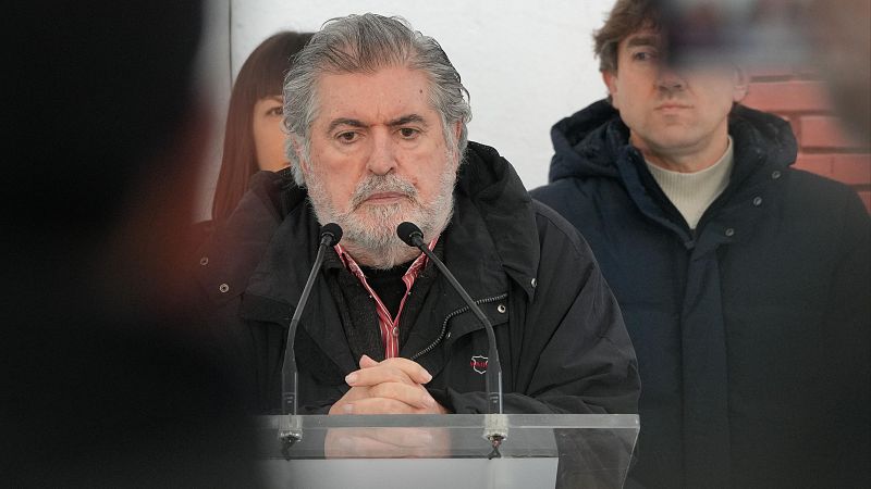 Jesús Eguiguren interviene serio tras un atril transparente con micrófonos en una rueda de prensa con fondo difuminado.