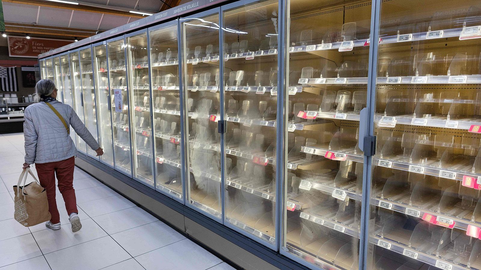 Una mujer camina junto a refrigeradores vacíos en un supermercado con estantes desiertos y suelo brillante.