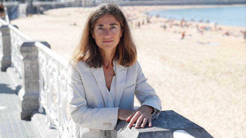 La política Pilar Zabala posa sonriente junto a la barandilla de un paseo marítimo en un día soleado, con una playa al fondo.