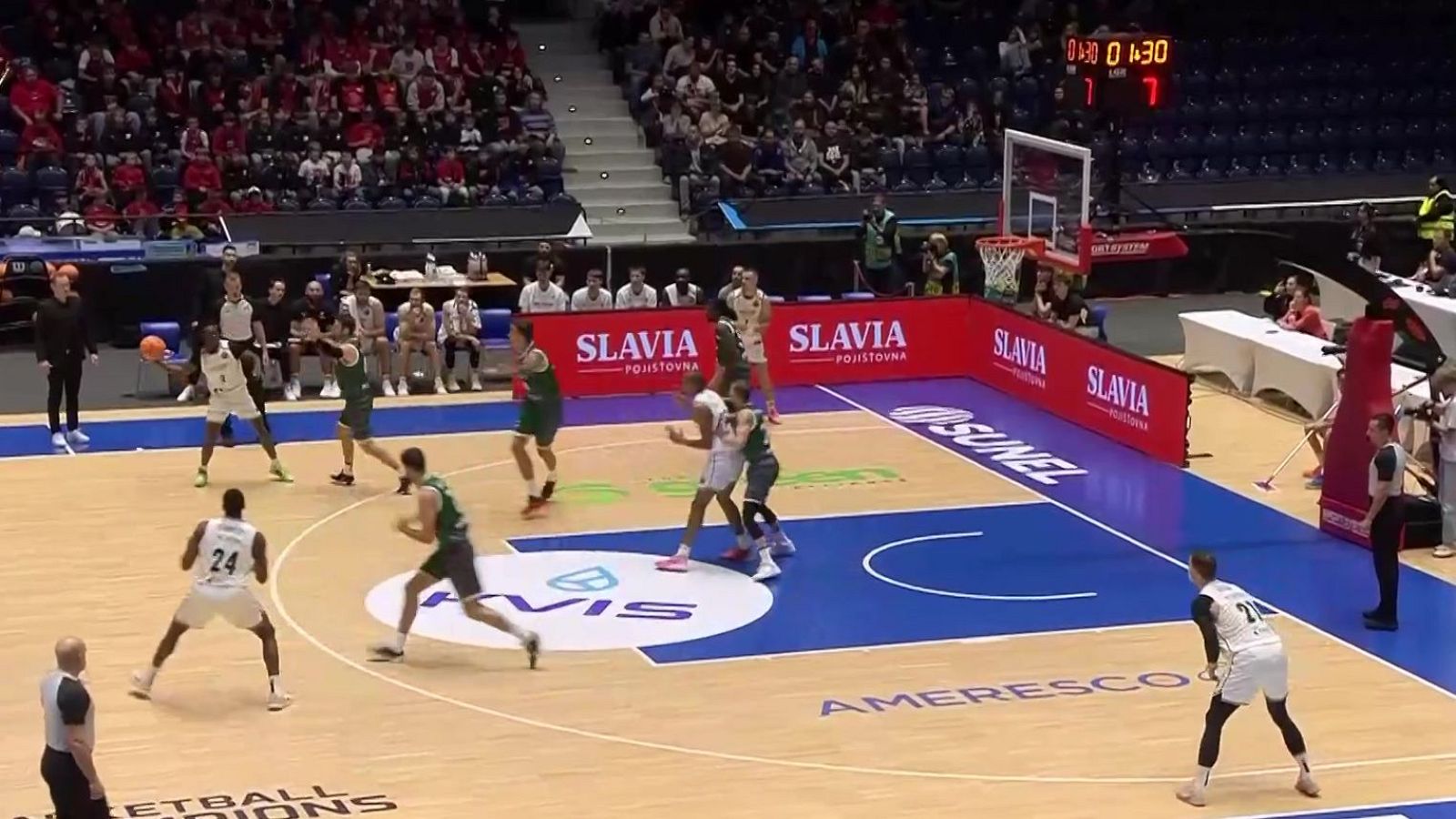 Plano general elevado de un partido de baloncesto en pabellón, con jugadores de blanco y verde disputando la posesión sobre el parqué de la cancha.