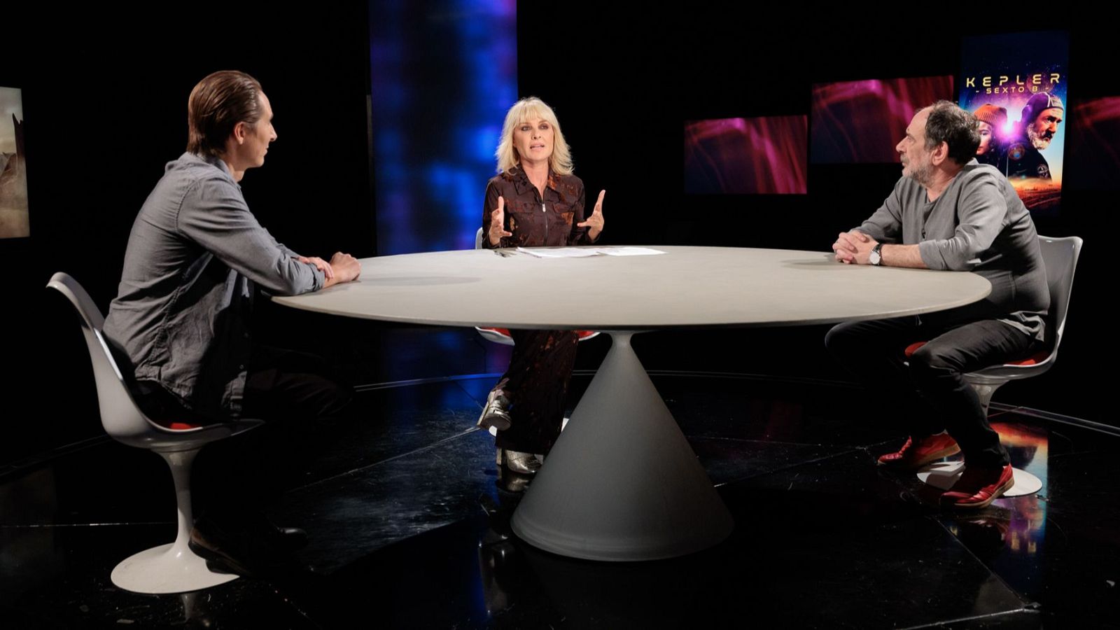 Tres personas conversan sentadas alrededor de una mesa circular en un plató de televisión con iluminación azul y magenta y suelo reflectante.