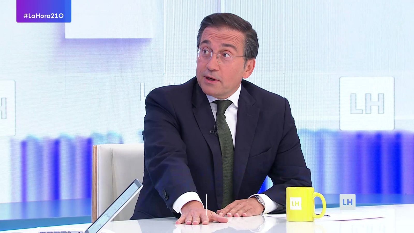 Plano medio de un hombre con traje azul y corbata verde oliva sentado en un plató de televisión, apoyado sobre una mesa con una taza amarilla.