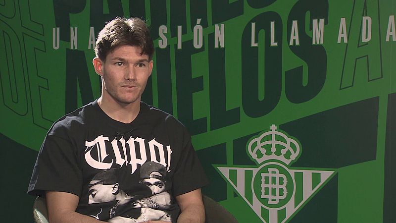 Joven futbolista con camiseta negra de Tupac durante una entrevista ante un panel corporativo con el escudo del Real Betis.