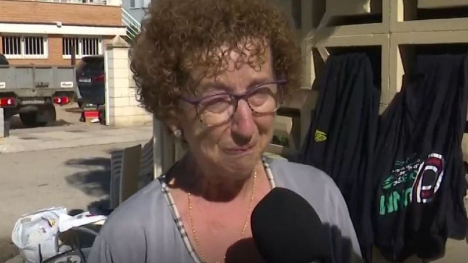 Mujer mayor de pelo rizado castaño y gafas moradas habla emocionada ante un micrófono de televisión en una calle residencial.