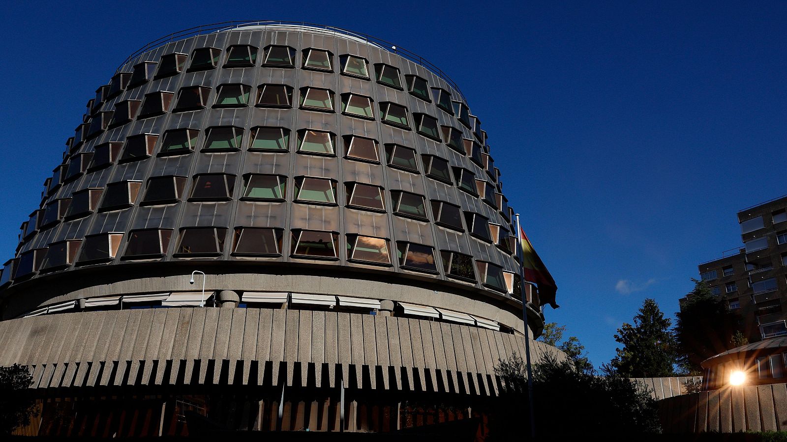 Vista exterior en contrapicado de la sede del Tribunal Constitucional de España en Madrid, destacando su arquitectura troncocónica escalonada y la bandera nacional.