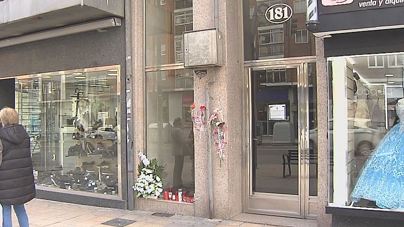 Entrada a un edificio residencial con un memorial de flores y velones en el suelo, flanqueada por escaparates comerciales.