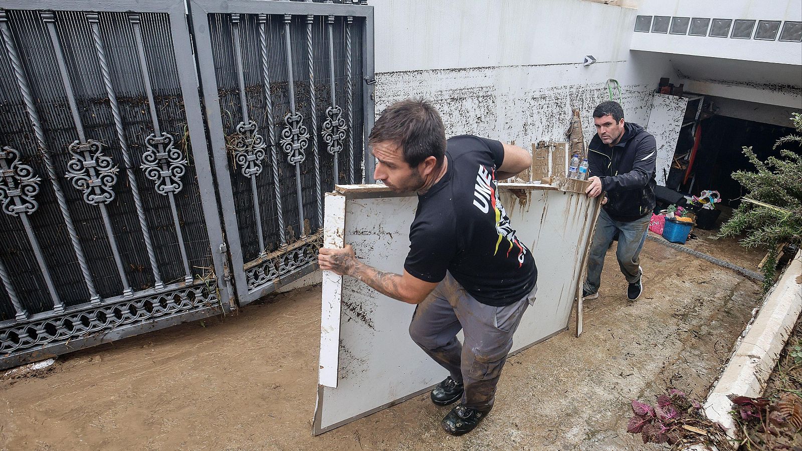 Dos hombres retiran un tablero embarrado en la rampa de un garaje inundado por una riada, uno de ellos con uniforme de la UME.