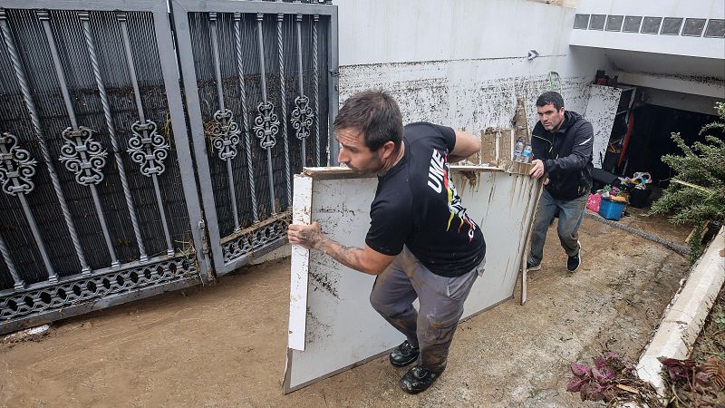 Dos hombres retiran un tablero embarrado en la rampa de un garaje inundado por una riada, uno de ellos con uniforme de la UME.