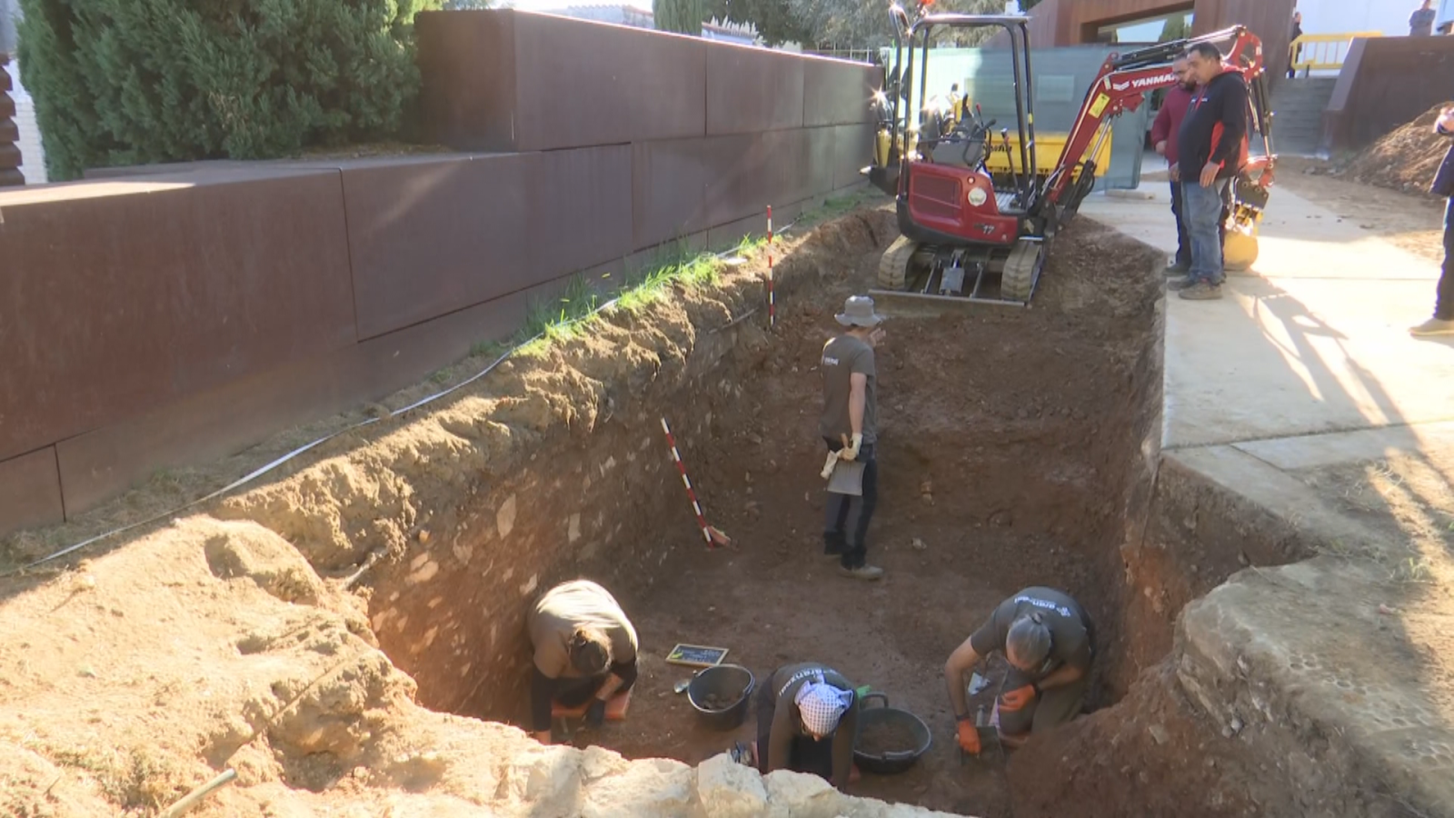 Vista picada de una excavación arqueológica en una fosa común con especialistas trabajando y una miniexcavadora al fondo.