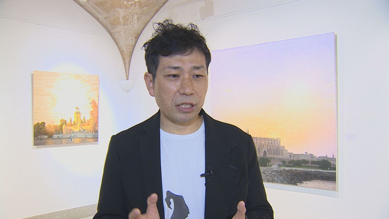 Hombre de rasgos asiáticos gesticula hablando en una galería de arte con cuadros iluminados al fondo.