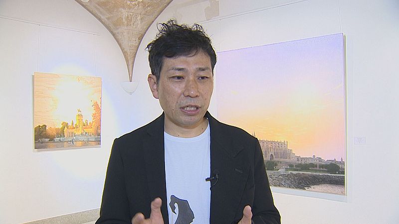 Hombre de rasgos asiáticos gesticula hablando en una galería de arte con cuadros iluminados al fondo.