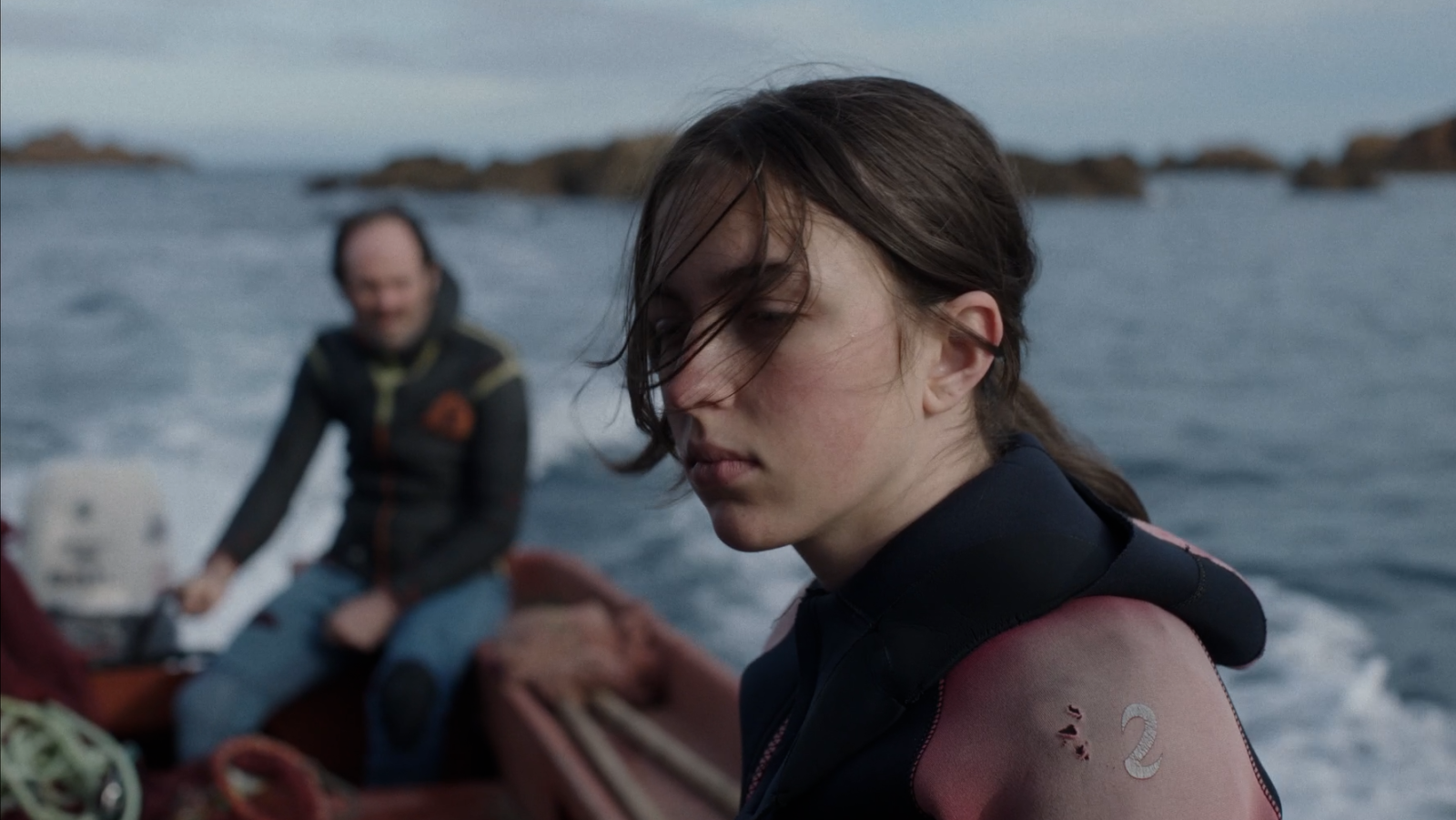 Joven con traje de neopreno y mirada seria a bordo de una barca en movimiento con un hombre al fondo y mar nublado.