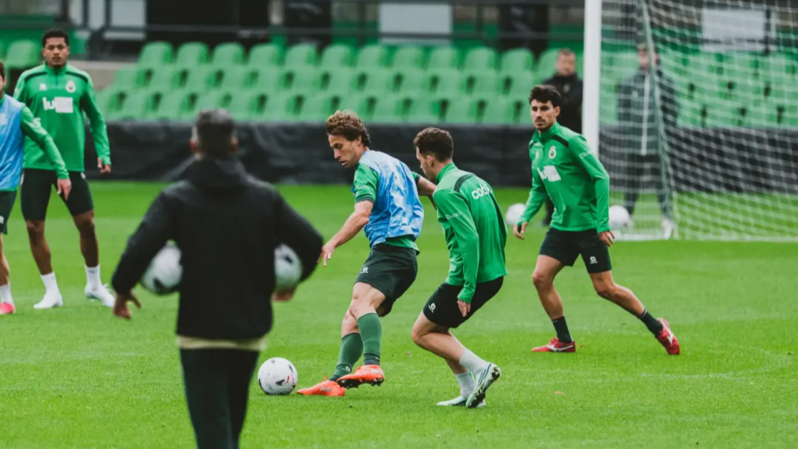 Futbolistas entrenando en un campo de césped natural, con un jugador conduciendo el balón ante la defensa de un compañero y un técnico observando.