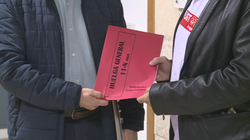 Dos personas sostienen una carpeta roja con el texto «HUELGA GENERAL» y «14-N 2012», destacando una acreditación sindical de UGT Aragón en el pecho.