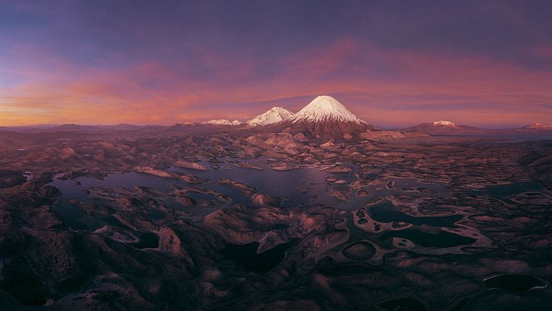 Impresionante paisaje volcánico de alta montaña al atardecer, con un volcán nevado al fondo y un laberinto de lagunas oscuras en primer plano.