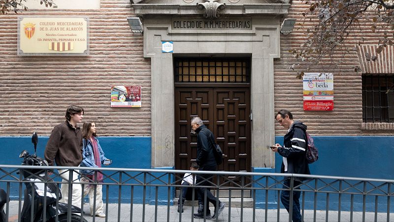 Fachada de ladrillo del Colegio Mercedarias de Don Juan de Alarcón con puerta de madera, placas informativas y peatones caminando por la acera.
