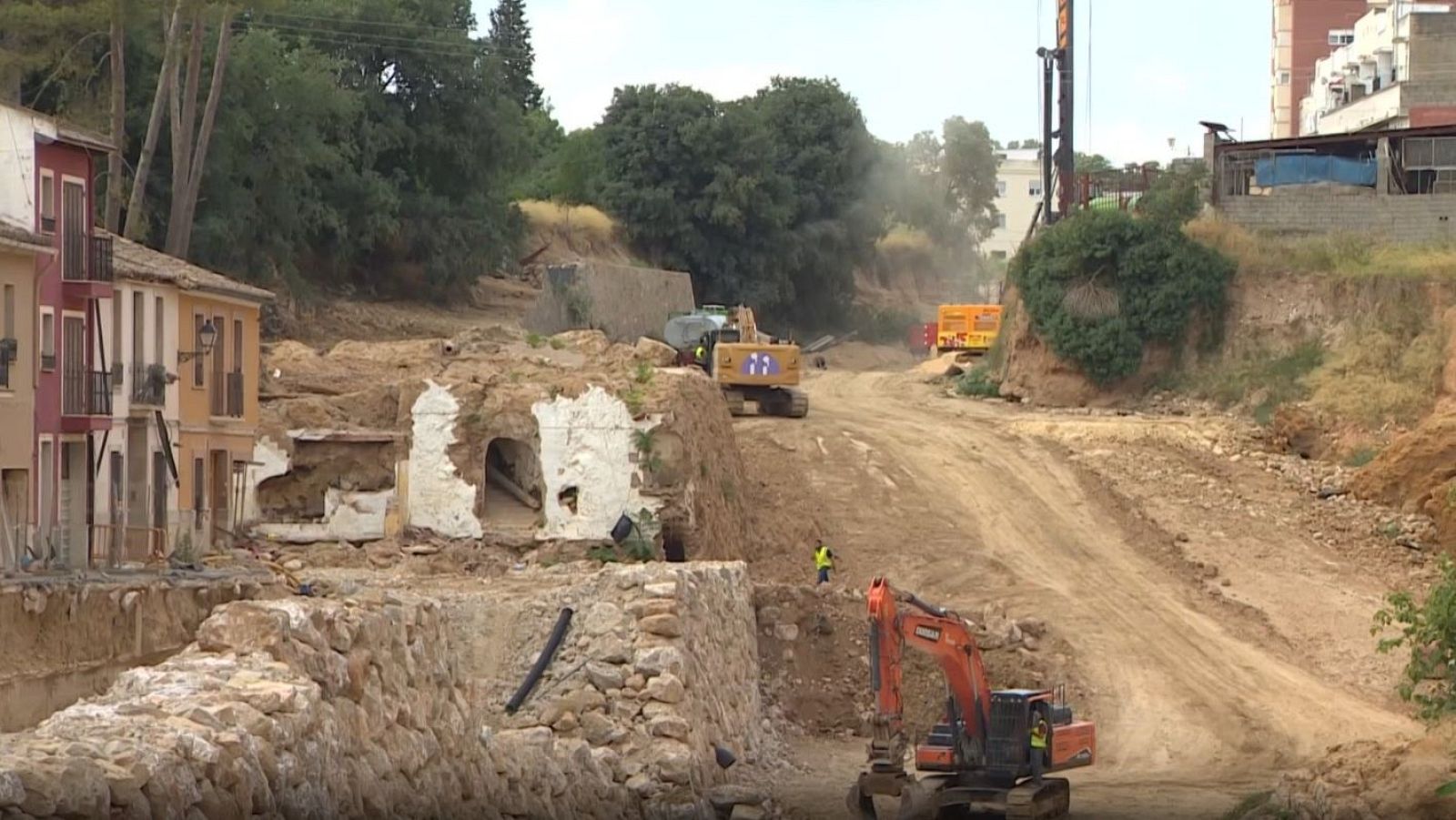 Zona de obras en una vaguada con excavadoras trabajando, operarios con chalecos reflectantes y viviendas cercanas bajo un cielo nublado.