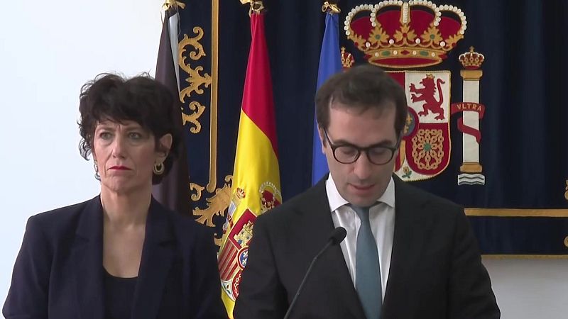 Carlos Cuerpo interviene ante un micrófono en un acto institucional junto a Cristina Pérez, con banderas oficiales y un repostero al fondo.