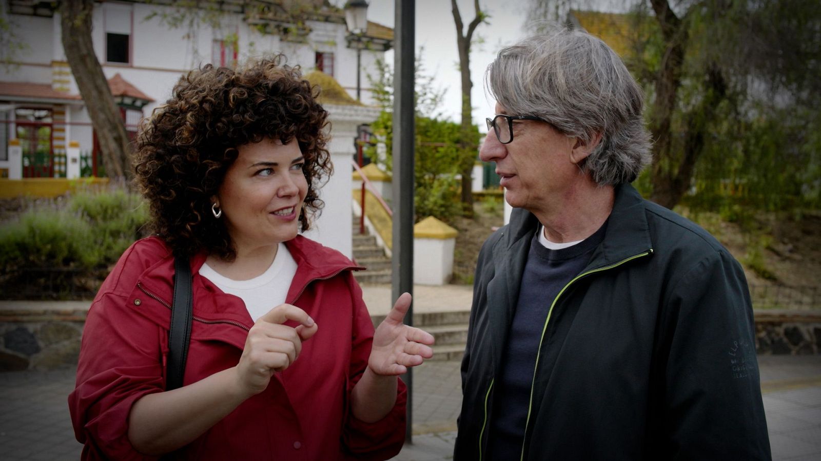 La mujer de pelo rizado y chaqueta roja gesticula sonriente mientras conversa al aire libre con el director y escritor David Trueba.