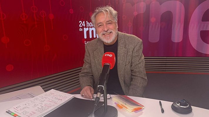 El escritor Manuel Rivas sonríe sentado ante un micrófono de RNE en un estudio con iluminación roja y notas sobre la mesa.
