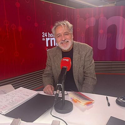El escritor Manuel Rivas sonríe sentado ante un micrófono de RNE en un estudio con iluminación roja y notas sobre la mesa.