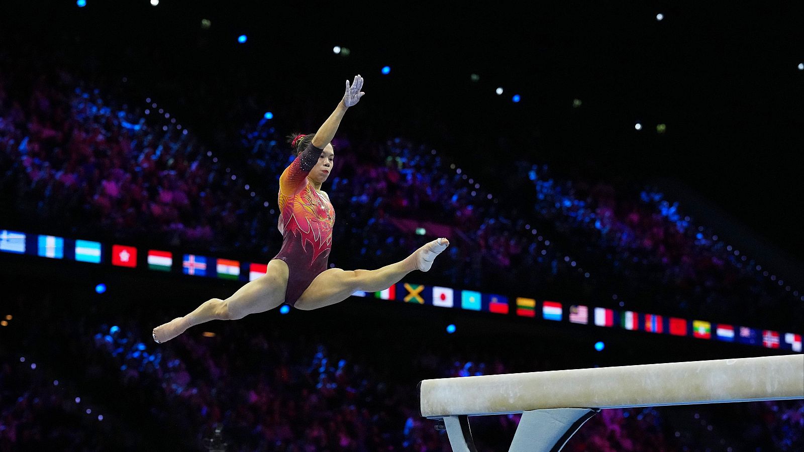 Gimnasta artística suspendida en el aire durante un salto en split sobre la barra de equilibrio, vistiendo maillot granate con detalles brillantes.