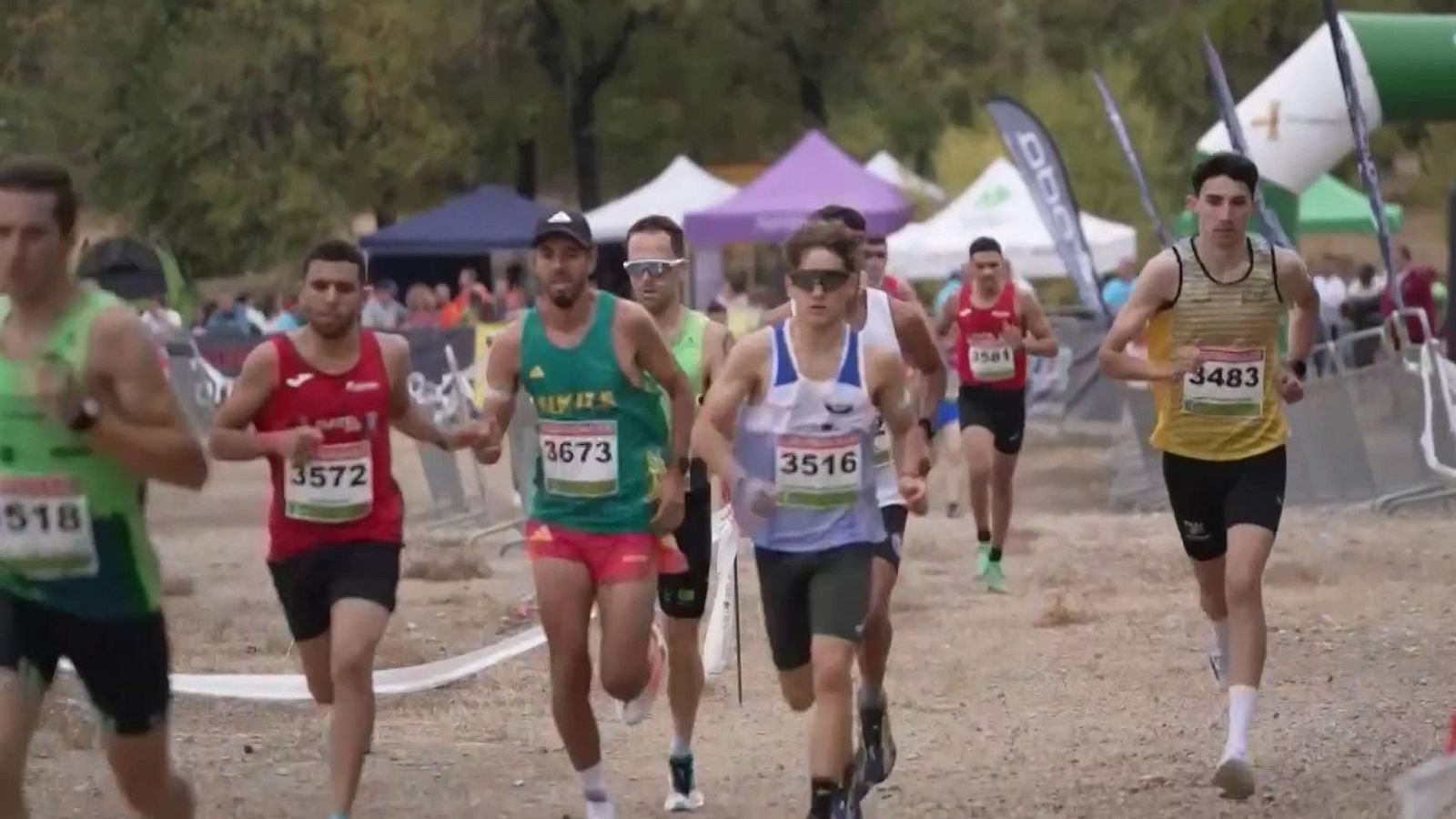 Grupo de atletas masculinos compitiendo en una carrera de campo a través por un sendero de tierra, mostrando dorsales y equipaciones deportivas variadas.