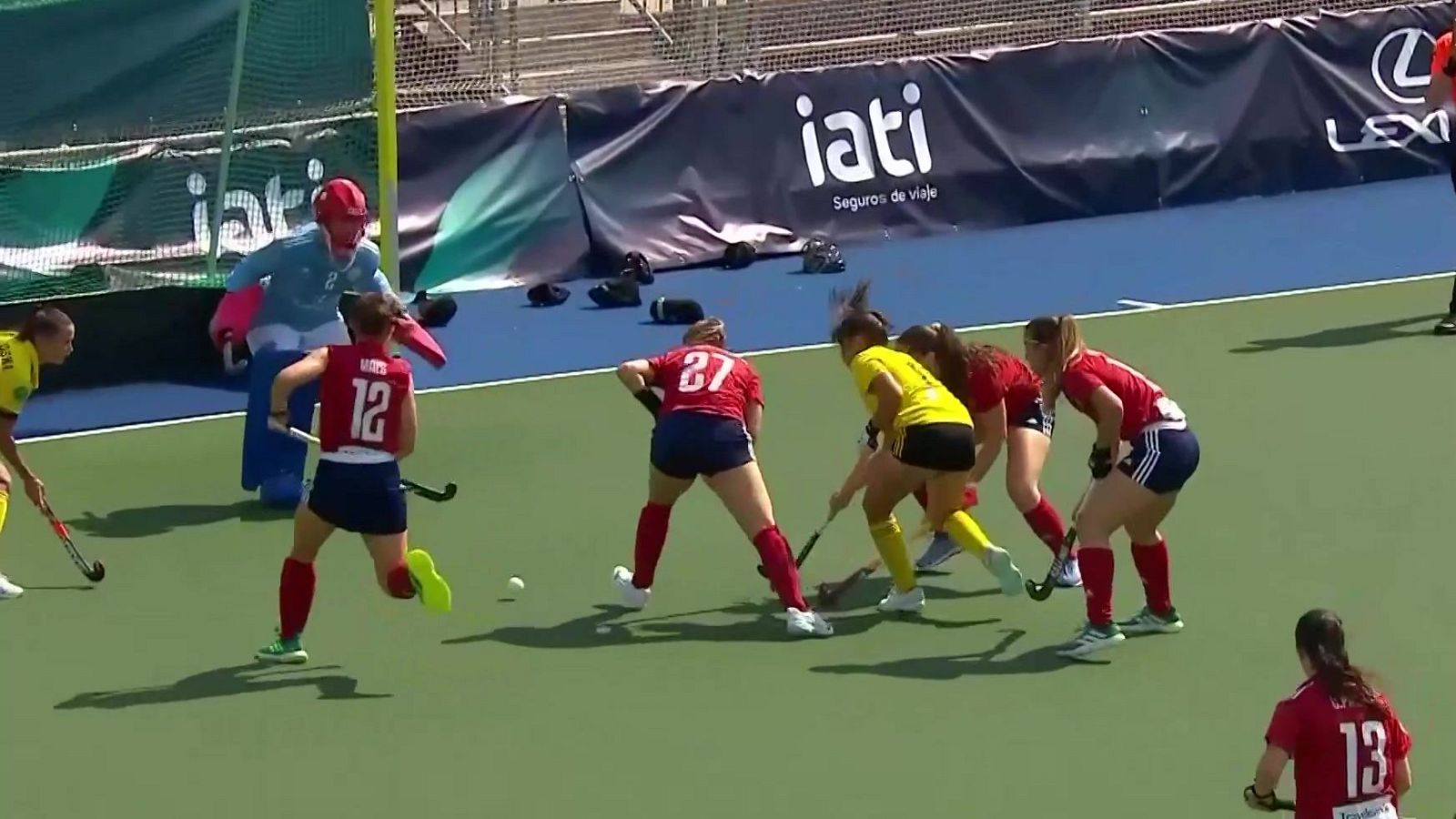 Jugadoras de hockey sobre hierba disputan un penalti córner en un campo de césped artificial verde con marcas azules, junto a una portería y vallas publicitarias.