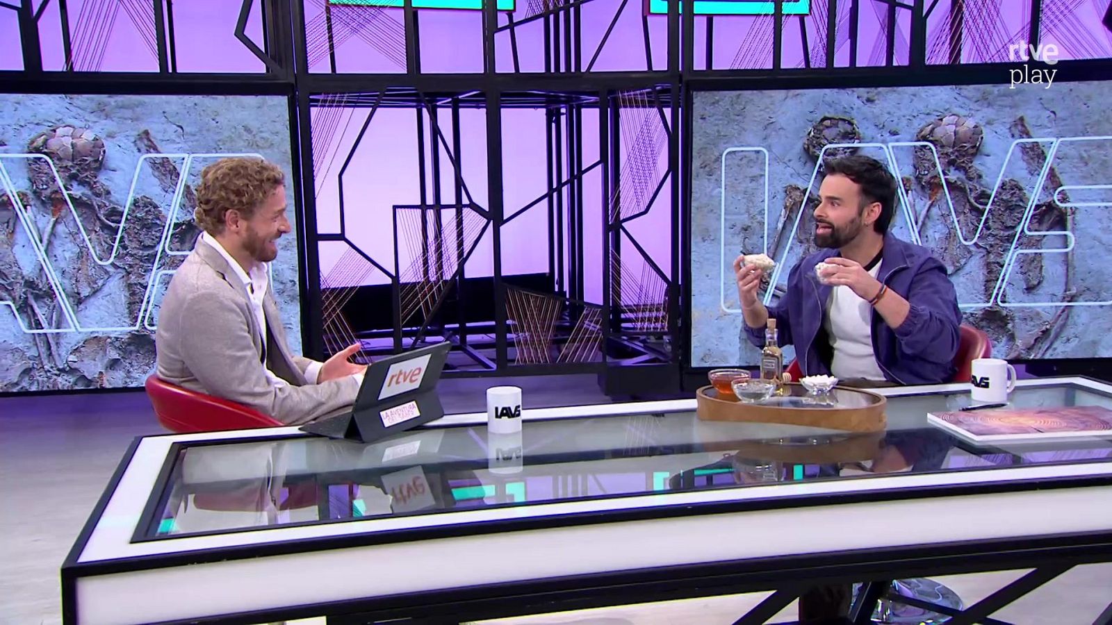 Dos hombres charlan en un plató de televisión frente a una mesa de cristal, rodeados de elementos de atrezo como tazas con logotipos, una bandeja con alimentos y pantallas que proyectan fósiles arqueológicos.
