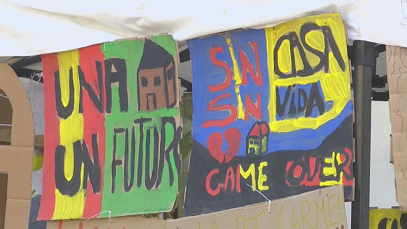 Carteles de protesta artesanales de cartón con los lemas «UNA CASA / UN FUTUR» y «SIN CASA / SIN VIDA» bajo una carpa blanca.