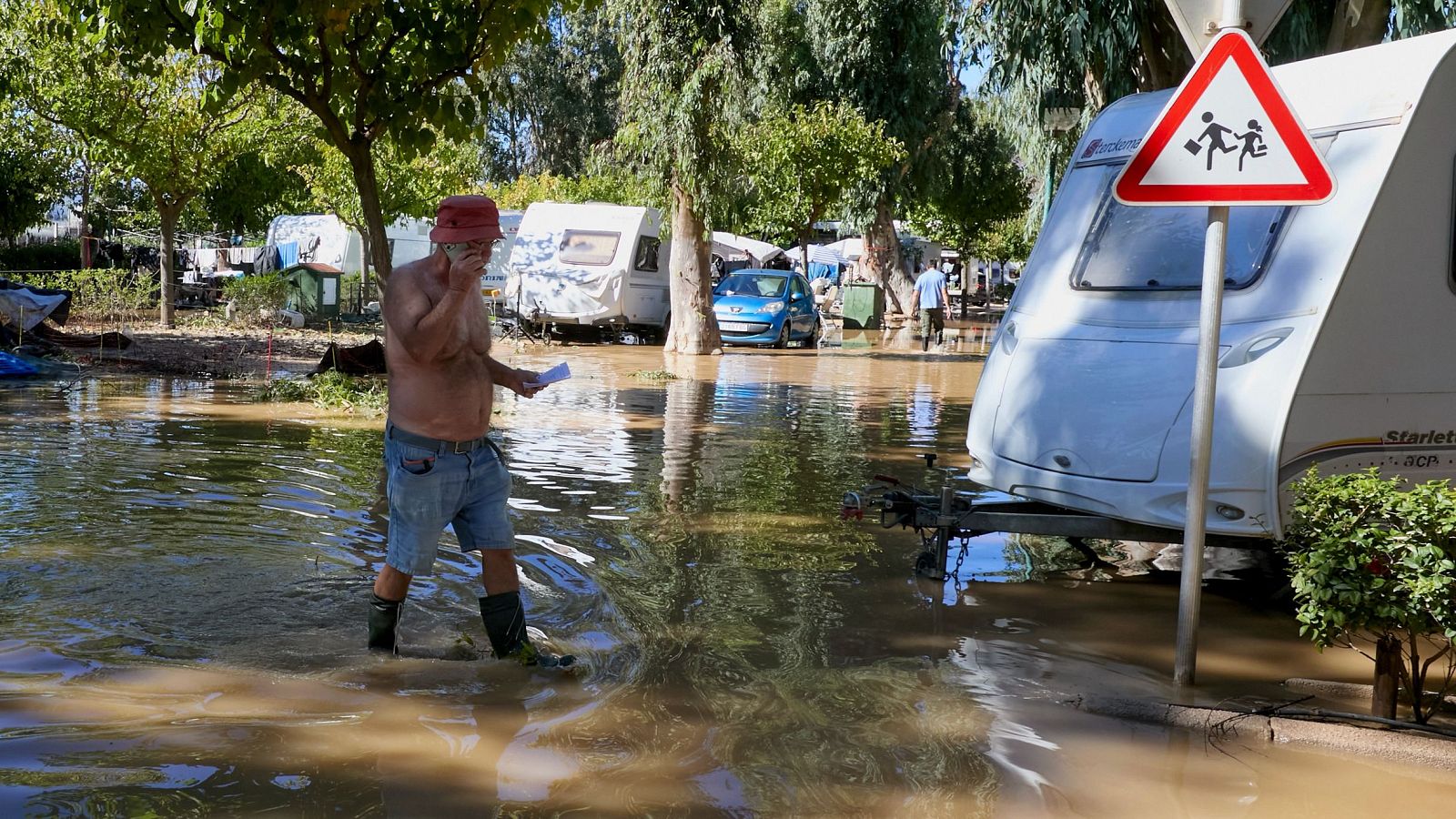 Un hombre sin camiseta y con botas de agua camina por una zona anegada tras una inundación, hablando por teléfono mientras al fondo se ven caravanas y árboles.