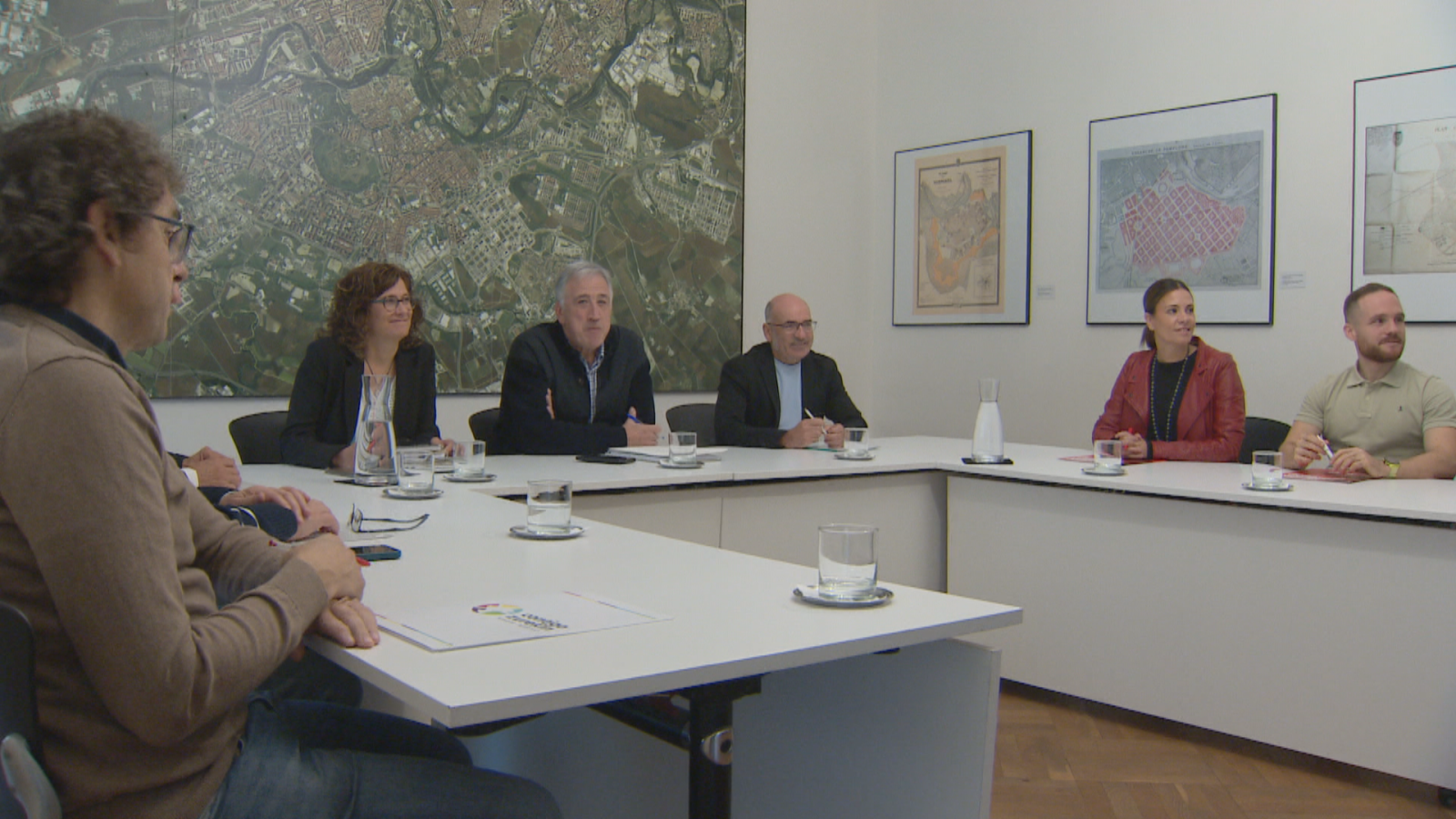 Reunión institucional en Pamplona entre EH Bildu, Contigo-Zurekin y PSN para negociar los presupuestos municipales en una sala con mesas en herradura y una ortofoto de la ciudad al fondo.