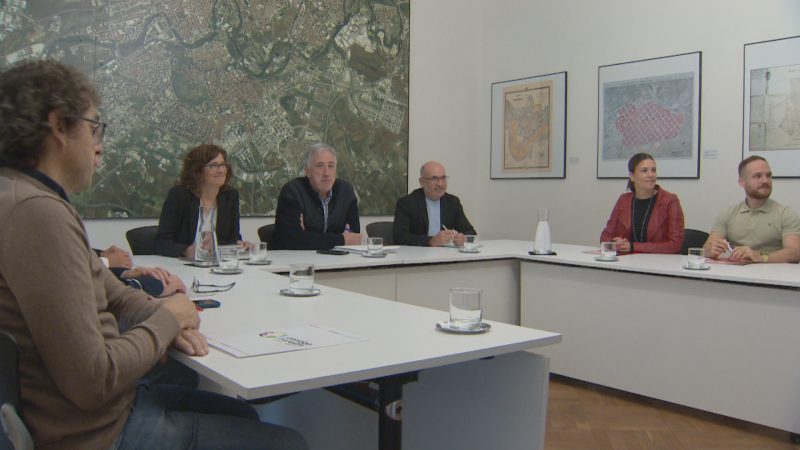 Reunión institucional en Pamplona entre EH Bildu, Contigo-Zurekin y PSN para negociar los presupuestos municipales en una sala con mesas en herradura y una ortofoto de la ciudad al fondo.