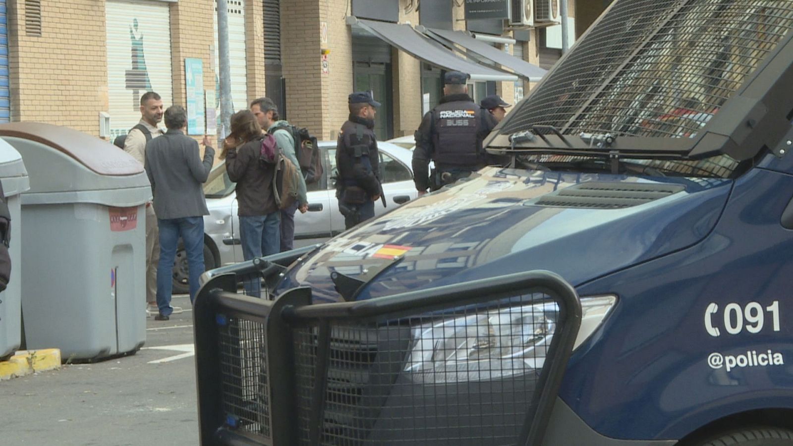 Furgón y agentes antidisturbios de la Policía Nacional desplegados en una calle urbana junto a un grupo de civiles.
