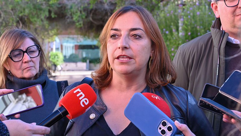 La política Miriam Andrés ofrece declaraciones a los medios en la calle, rodeada de micrófonos y periodistas.